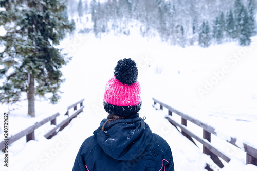 Girl in winter warm jacket and pink woolen hat walking in snowy winter forest