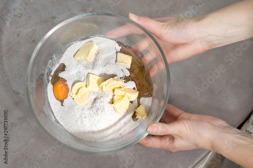 Kobiece dłonie trzymają misę z niewymieszanymi składnikami do wypieku ciasta w szklanej misce. Jajka, mąka, margaryna w szklanej misce trzymana przez kobiece dłonie.