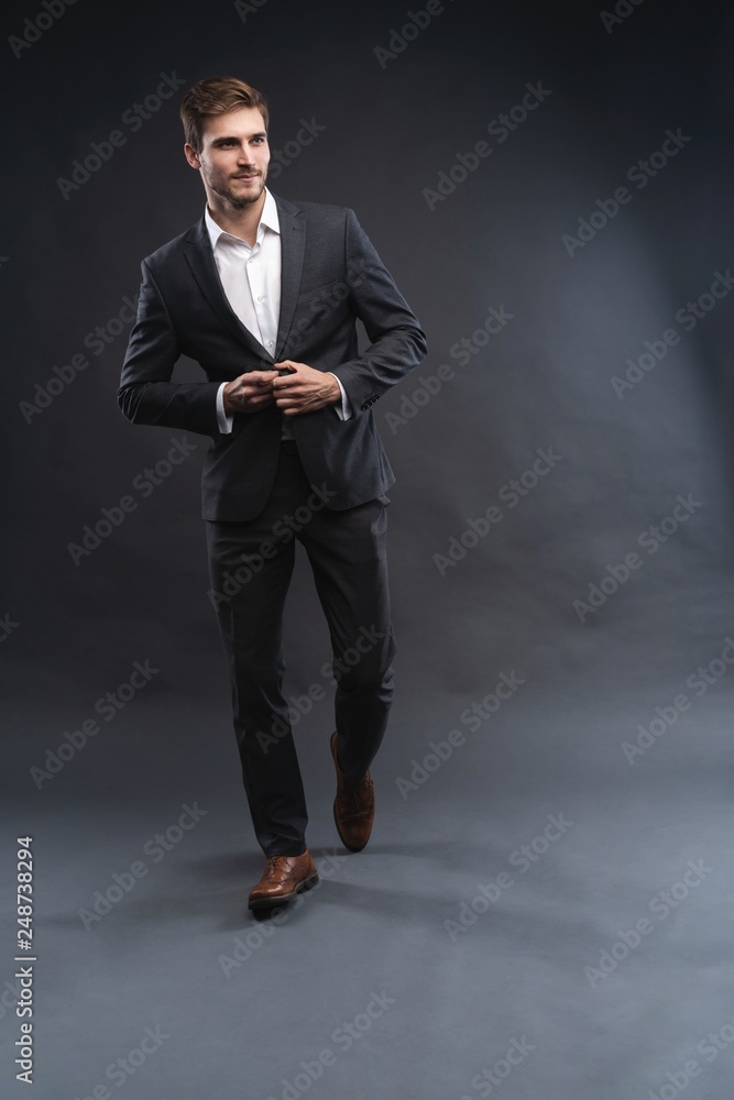 Full length portrait of a handsome young businessman dressed in suit isolated over gray background, posing, looking away, walking Full length portrait of a handsome young businessman dressed in suit isolated over gray background, posing, looking away, walking