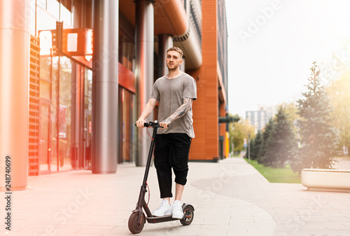 Attractive young man riding a kick scooter at cityscape background.