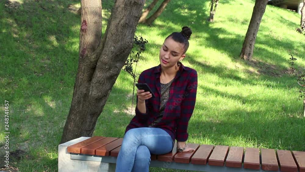Young beautiful girl in a plaid shirt is sitting on a bench in the park, smiling and writing a message on the internet chatting on her smartphone. Young beautiful girl in a plaid shirt is sitting on a bench in the park, smiling and writing a message on the internet chatting on her smartphone.