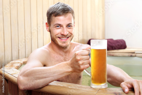 Piwne spa. Przystojny mężczyzna zażywa kąpieli w bali z woda termalną pijąc piwo kuflowe.