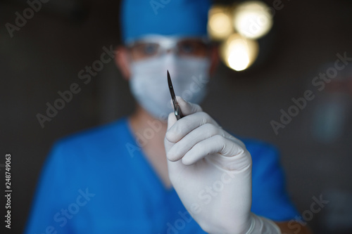 Fototapeta Professional surgeon in blue uniform in a medical mask in sterile gloves holds a metal scalpel in his hand