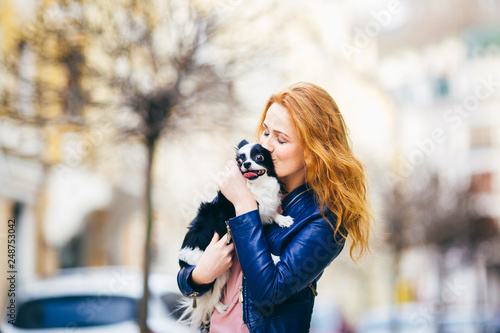 A young redhaired Caucasian woman with freckles holds and kisses, embracing black and white shaggy dog of Chihuahua breed. girl dressed in blue leather jacket, stands on street in spring in Europe © Elizaveta