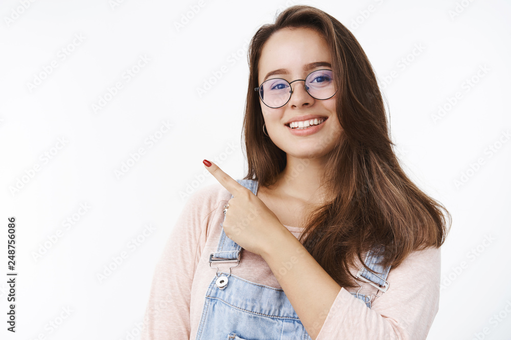 Joyful pleasant young female assistant in glasses and overalls smiling broadly pointing at upper left corner showing awesome place to hang out, asking click on copy space over gray background Joyful pleasant young female assistant in glasses and overalls smiling broadly pointing at upper left corner showing awesome place to hang out, asking click on copy space over gray background
