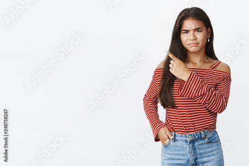 Moody and displeased immature teenage girl complaining making upset and dissatisfied grimace, wrinkling nose and frowning from dislike as pointing at upper left corner making negative opinion