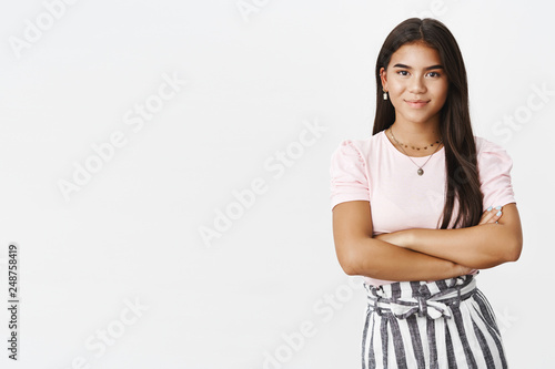 Studio shot of assertive and self-assured good-looking young adolescent girl in skirt and blouse holding hands crossed over chest in confident and ready pose, smiling friendly at camera