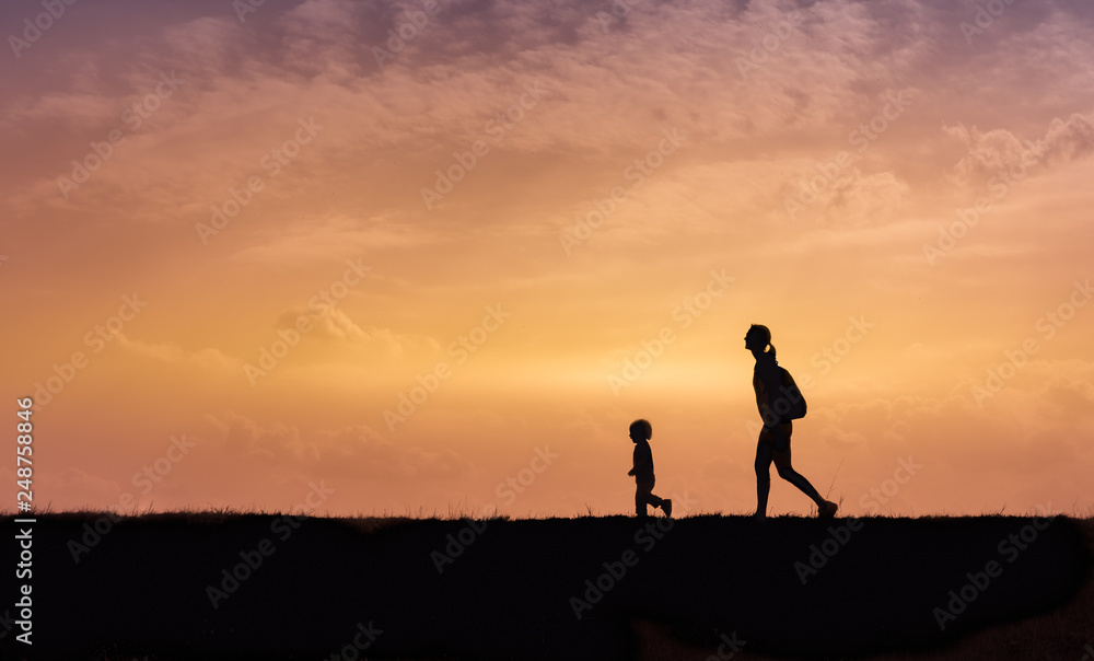 Silhouette of mother and her little child walking together outdoors at sunset.