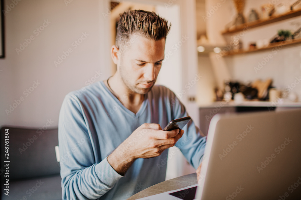 Handsome coworker man working at living room at home. Man sitting at wooden table using laptop and mobile phone. Blurred background Handsome coworker man working at living room at home. Man sitting at wooden table using laptop and mobile phone. Blurred background
