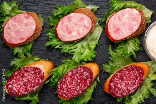 Several sandwiches with sausage and salami and sauce on a black board, flat lay