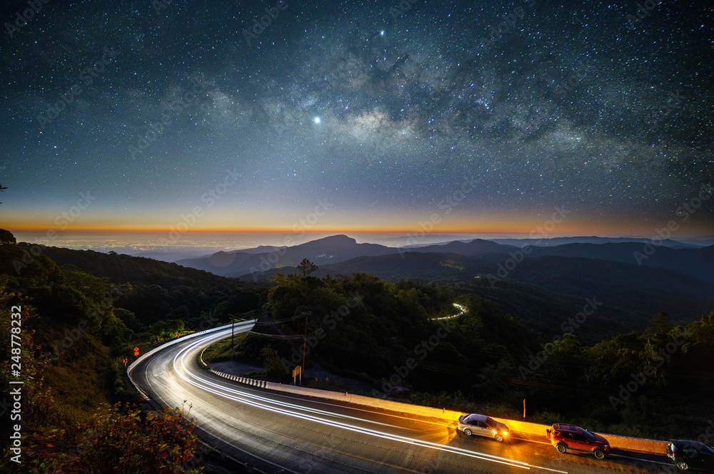 Milky way and stars in the night with car light trails on the mountains