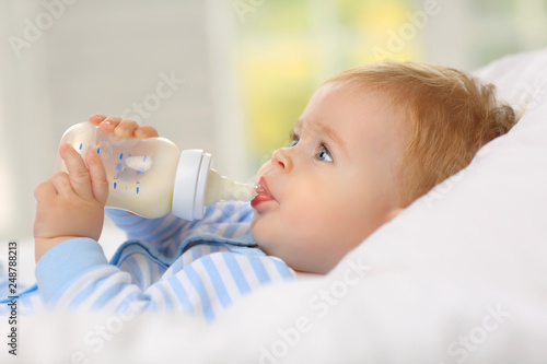 Billede på lærred Charming blue-eyed baby 7 month old lies in bed and drinks milk from a bottle