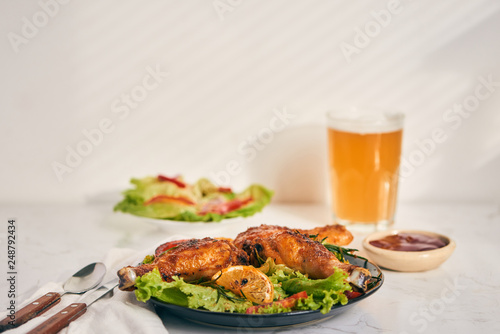 Grilled chicken legs roasted on the grill on dark plate with tomato sauce in a bowl and lettuce leaves, glass mug of beer