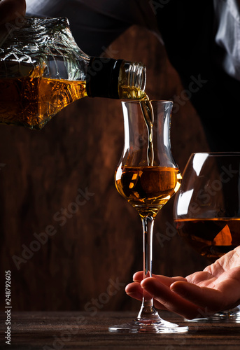 The bartender pours the grappa or liquor in small shot glass on the old bar counter. Vintage wooden background in pub or bar, night mood. Place for text, toning, selective focus photo