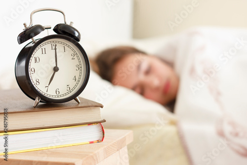 Teenager girl sleeping in a white bed. Alarm clock in the foreground on a pile of books
