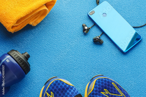 Sports equipment flat lay composition, running shoes, yellow towel, smartphone, earphones, fitness tracker and bottle of water on blue background. Concept healthy lifestyle, sport and diet.