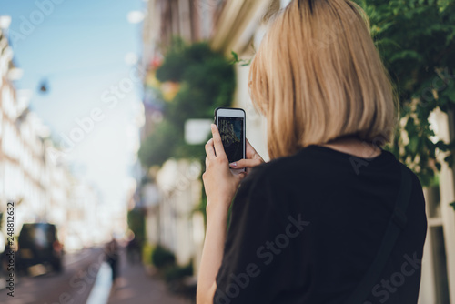 Back view of teenage hipster girl taking picture of the streets on her mobile phone camera while travelling around Europe, using modern cell telephone to take photos, copy space for your text or logo