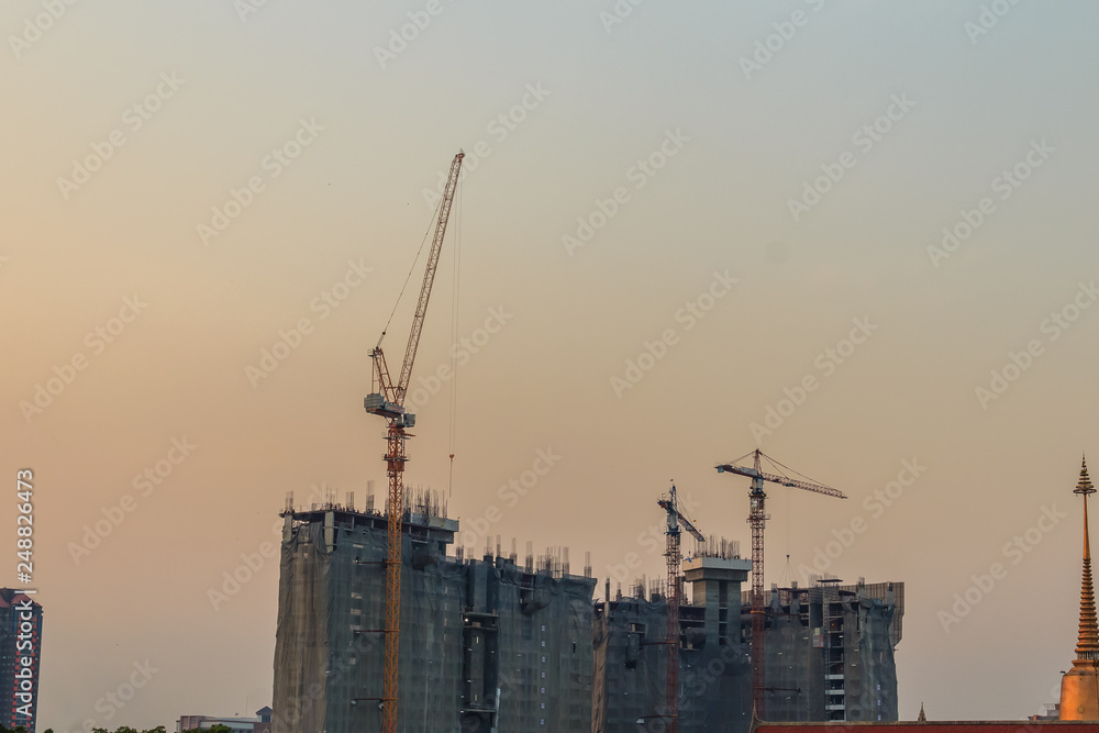 Luffing jib tower crane at condominium construction site over steel framework among high rise building. Skyscraper building construction with the tower cranes on top under the dramatic sky background.