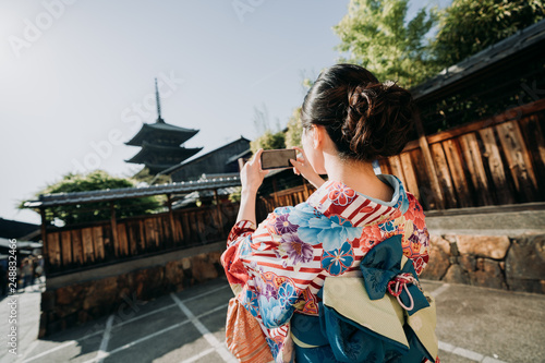 back view of young asian female japanese using cellphone taking picture of yasaka pagoda kyoto japan under sunset. girl in colorful kimono cloth hold mobile phone photographing in sannen zaka street