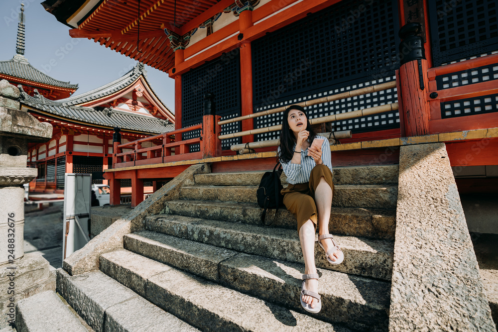 cheerful girl tourist checking information on line in smart phone sitting on stairs of japanese red wooden shinto. young woman backpacker visit kiyomizu dera 
temple kyoto japan in summer.