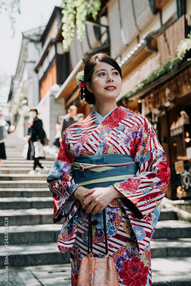 local japanese woman standing on stairs in sannen zaka street wearing long colorful kimono dress. young girl in traditional cloth smiling curious sightseeing vendor store shop in background.