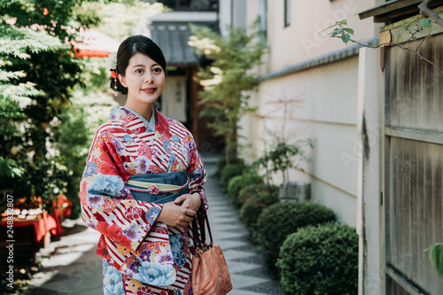 local japanese woman smiling standing outdoor of her house in front garden with trees and plants near bamboo door. young girl wearing flower colorful kimono dress under sunlight waiting.