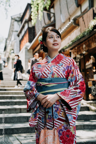 local japanese woman standing on stairs in sannen zaka street wearing long colorful kimono dress. young girl in traditional cloth smiling curious sightseeing vendor store shop in background.