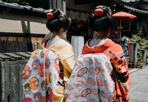 back view of two maiko geisha walking on a street of Gion in Kyoto Japan. japanese girls wearing traditional cloth dress kimono with amazing makeup and hair in old town on sunny day.