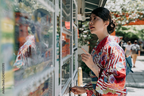 thirsty japanese lady in kimono dress in hot summer insert coins to vendor machine selling snck and drinks. young girl in traditional cloth choosing buying water with finger pointing on sunny day. photo