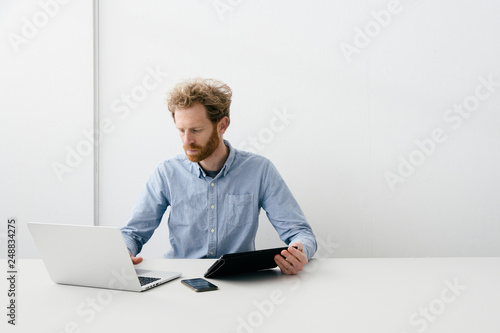 Thirty something bearded man looking thoughtfully on his laptop, phone and tablet at hand, sitting at an empty light colored desk.