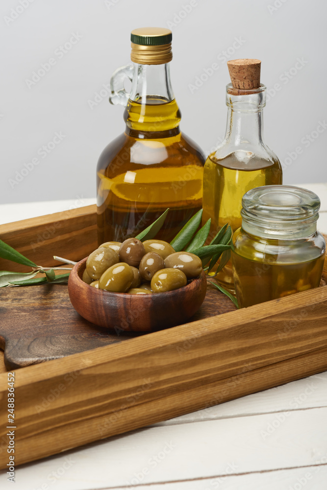 wooden tray with bowl of olives, oil bottles and olive tree leaves on white surface wooden tray with bowl of olives, oil bottles and olive tree leaves on white surface