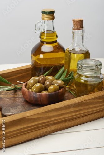 wooden tray with bowl of olives, oil bottles and olive tree leaves on white surface