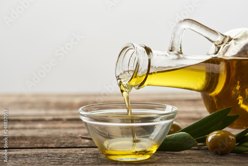 pouring oil from bottle into glass bowl, olive tree leaves and olives on wooden surface