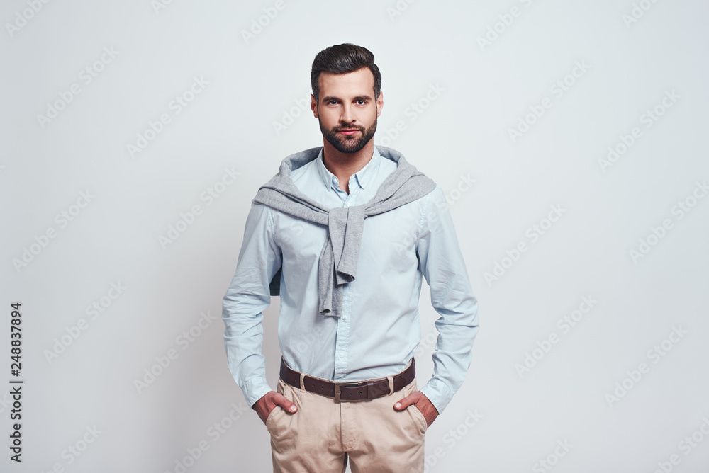 Fototapeta premium Irresistible man. Bearded young man is looking at camera, keeping hands in his pockets while standing against grey background