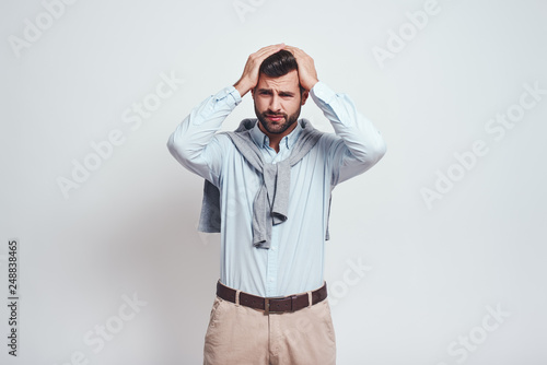 Emotional stress. Close-up portrait of confused young man keeping head in his hands and making a sad emotion while standing against grey background.