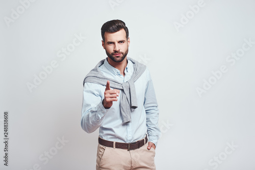 Who are you? Close-up studio portrait of serious handsome man pointing his finger and looking at camera while standing against grey background © Friends Stock