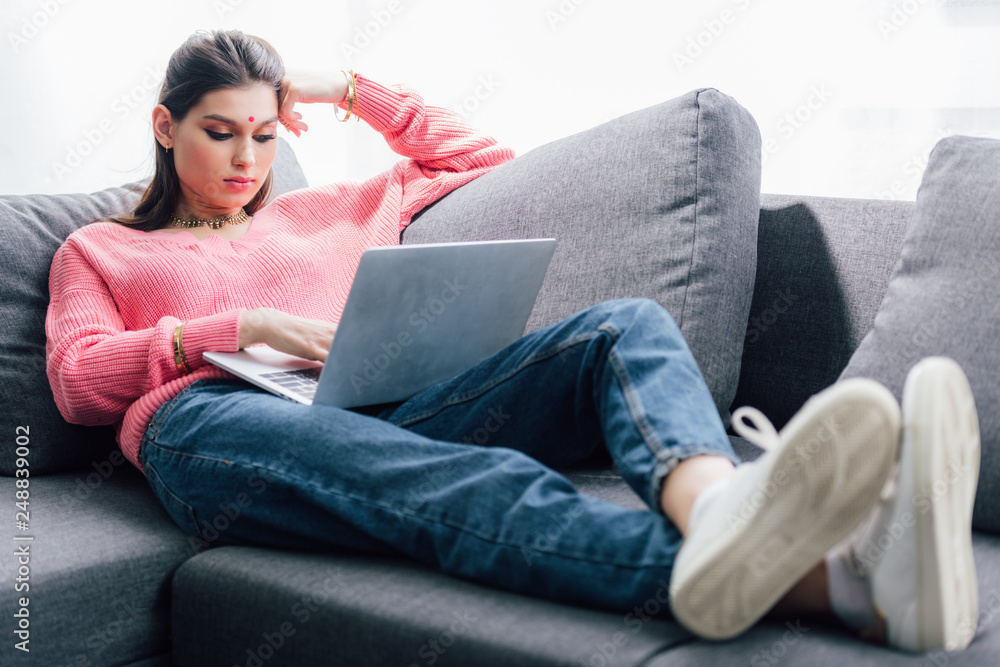 beautiful indian woman with bindi using laptop while lying on sofa beautiful indian woman with bindi using laptop while lying on sofa