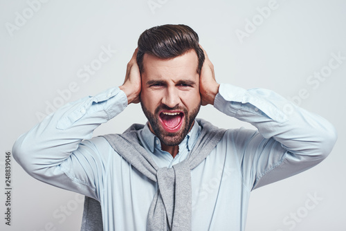 Bla bla bla! Young handsome man is stressed by noise, closing ears with both hands while standing on a grey background