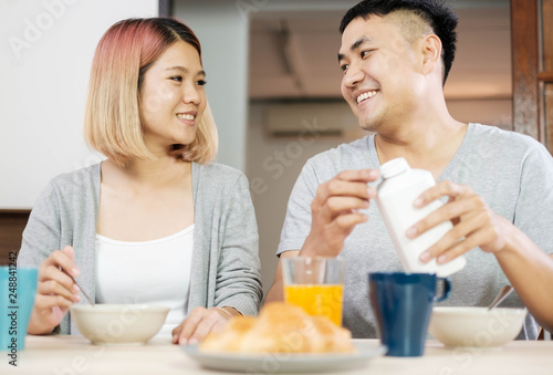 happy asian couple in pajamas sitting at table in kitchen at home in morning and having cereal breakfast together.husband pour milk into wife bowl.