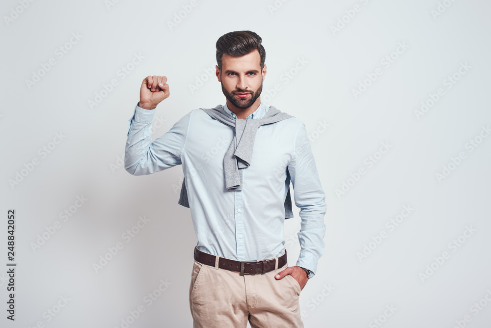 You can do it! Young attractive brunet in blue shirt is raising one hand clanched in fist above his head while standing against grey background You can do it! Young attractive brunet in blue shirt is raising one hand clanched in fist above his head while standing against grey background
