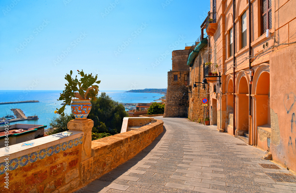 Old narrow street with Ceramic pot with cactus on wall and view on the port and sea in front of Sciacca, Sicily, Italy