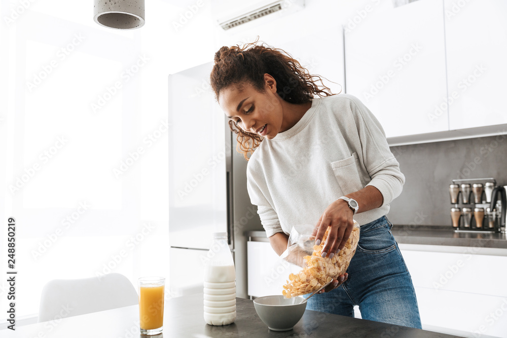 Image of smiling african american woman making breakfast with corn flakes and milk, in stylish kitchen - obrazy, fototapety, plakaty 