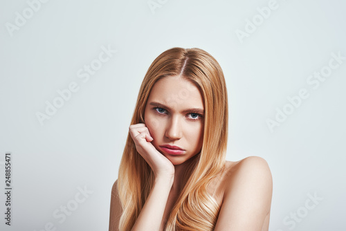 Bored. Upset young blonde woman looking at camera and making a sad face while standing in studio on a grey background