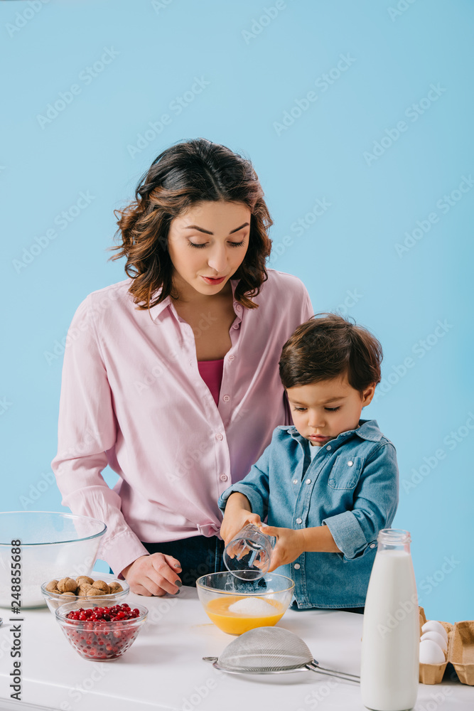 cute little boy adding sugar to whipped eggs while mommy looking at him isolated on blue cute little boy adding sugar to whipped eggs while mommy looking at him isolated on blue