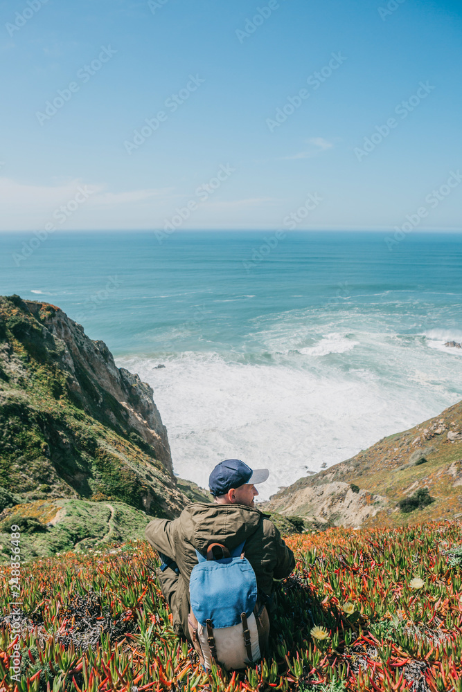 A tourist man with a backpack sits in solitude among flowers and plants on a hill and enjoys a beautiful view of the Atlantic Ocean on a spring day.