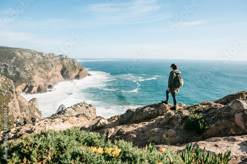 A tourist with a backpack on top of a cliff admires a beautiful view of the Atlantic Ocean in Portugal.
