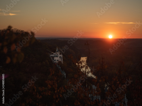 Sunset in Ukraine. Monastery on hills in evening. Narrow river flowing below monastery. Autumn nature landscape. Colorful dense forest on hills in October. Blurred background. Selective soft focus