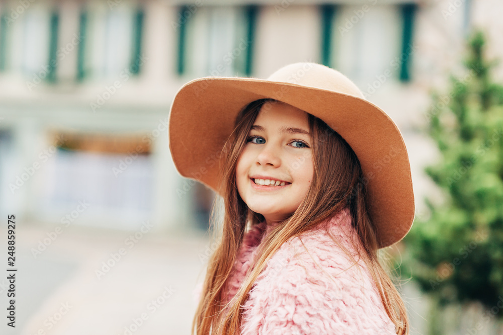 Outdoor portrait of pretty young kid girl wearing brown hat and pink coat, street fashion for children