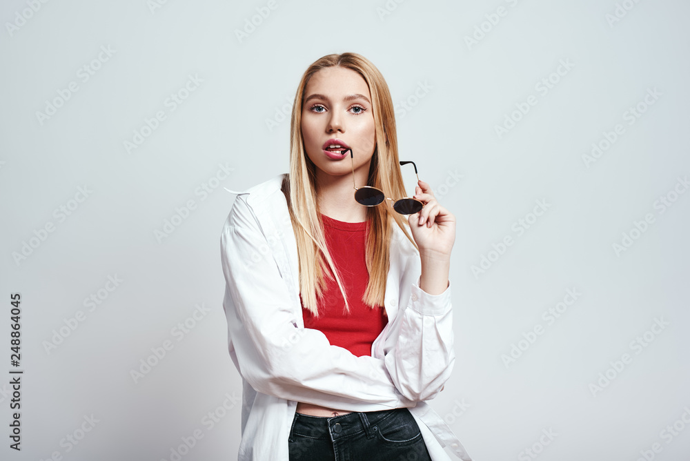 In her own style. Studio shot of pretty blonde woman holding sunglasses and looking at camera with open mouth while standing in studio against grey background In her own style. Studio shot of pretty blonde woman holding sunglasses and looking at camera with open mouth while standing in studio against grey background