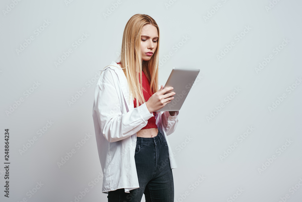 Very busy. Serious blonde girl in a casual wear is using her digital tablet for study while standing on a grey background. Digital concept Very busy. Serious blonde girl in a casual wear is using her digital tablet for study while standing on a grey background. Digital concept